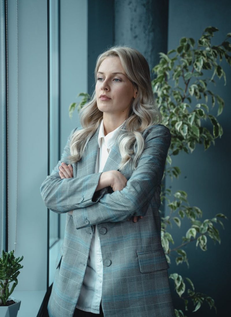 woman in gray blazer standing near green plant with a serious look