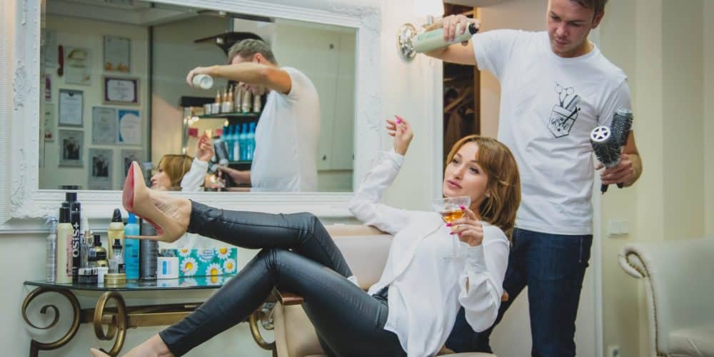 woman sitting on the salon chair while holding vodka glass and man at her back white spraying her hair